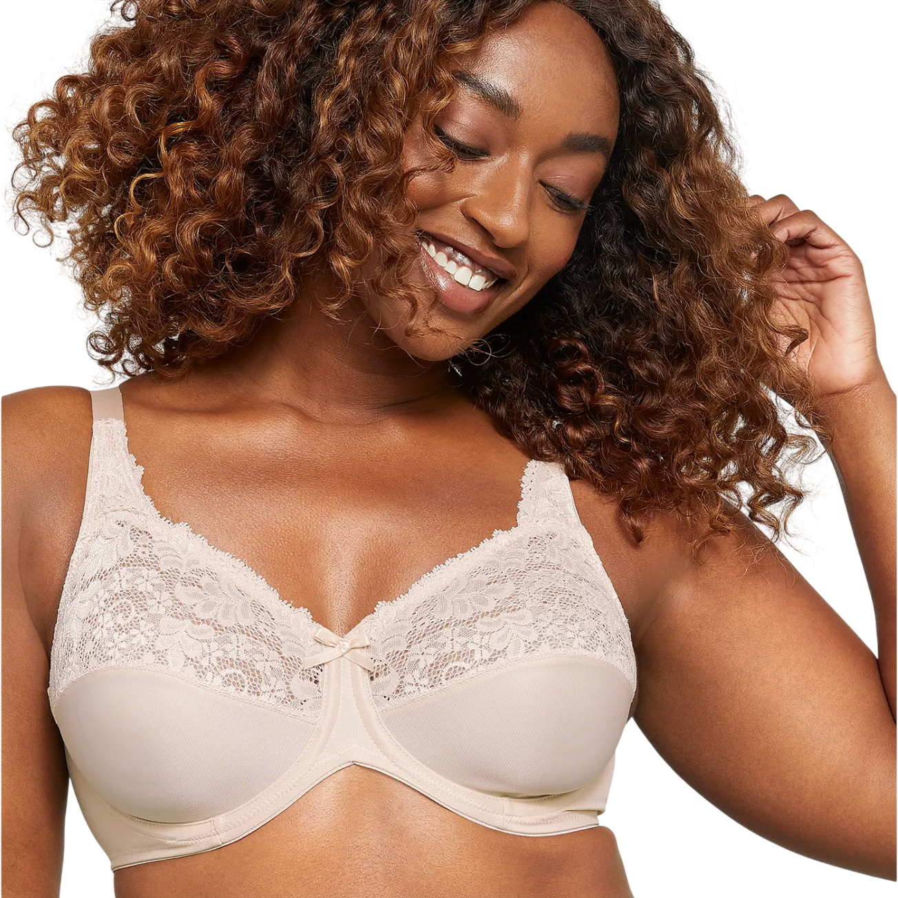 A woman is wearing a beige lace bra with intricate floral details and underwire support, smiling and touching her curly hair.