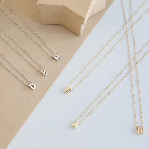 Five gold-plated initial necklaces with letters 'A,' 'B,' 'D,' 'N,' and 'R' hang on thin chains, displayed against a blue and white background.
