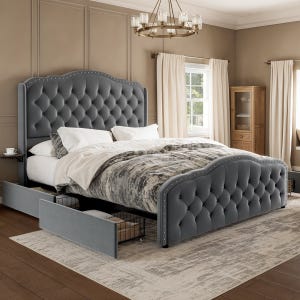 A tufted gray bed frame with a scalloped headboard and footboard, featuring nailhead trim and built-in storage drawers beneath. It is placed in a classic-style bedroom with beige walls, cream curtains, and a wooden cabinet.