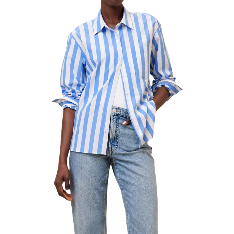 A model wears a blue and white vertically striped poplin shirt with a collared design and long sleeves, paired with light blue jeans. The shirt is partially tucked in and has a casual, slightly oversized fit.