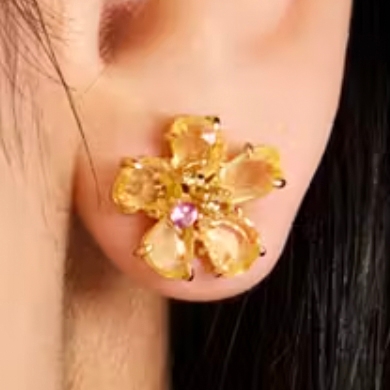 A flower-shaped earring with amber petals and a central pink gemstone, attached to an earlobe.