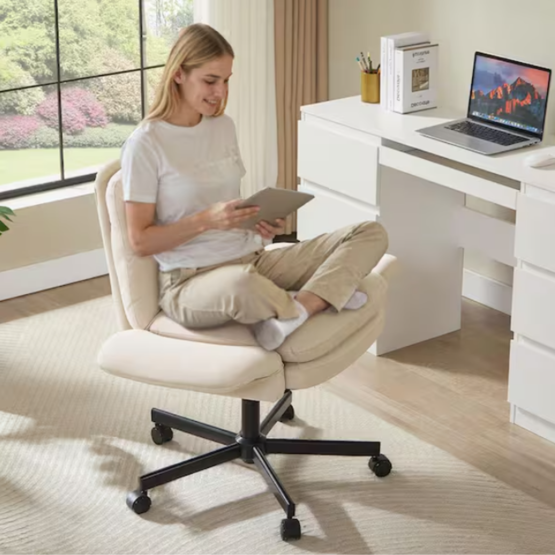 A person sits cross-legged on an adjustable office chair with a cushioned seat and backrest, holding a tablet near a sleek white desk with a laptop, books, and a pencil holder. The room has large windows and light-colored decor.