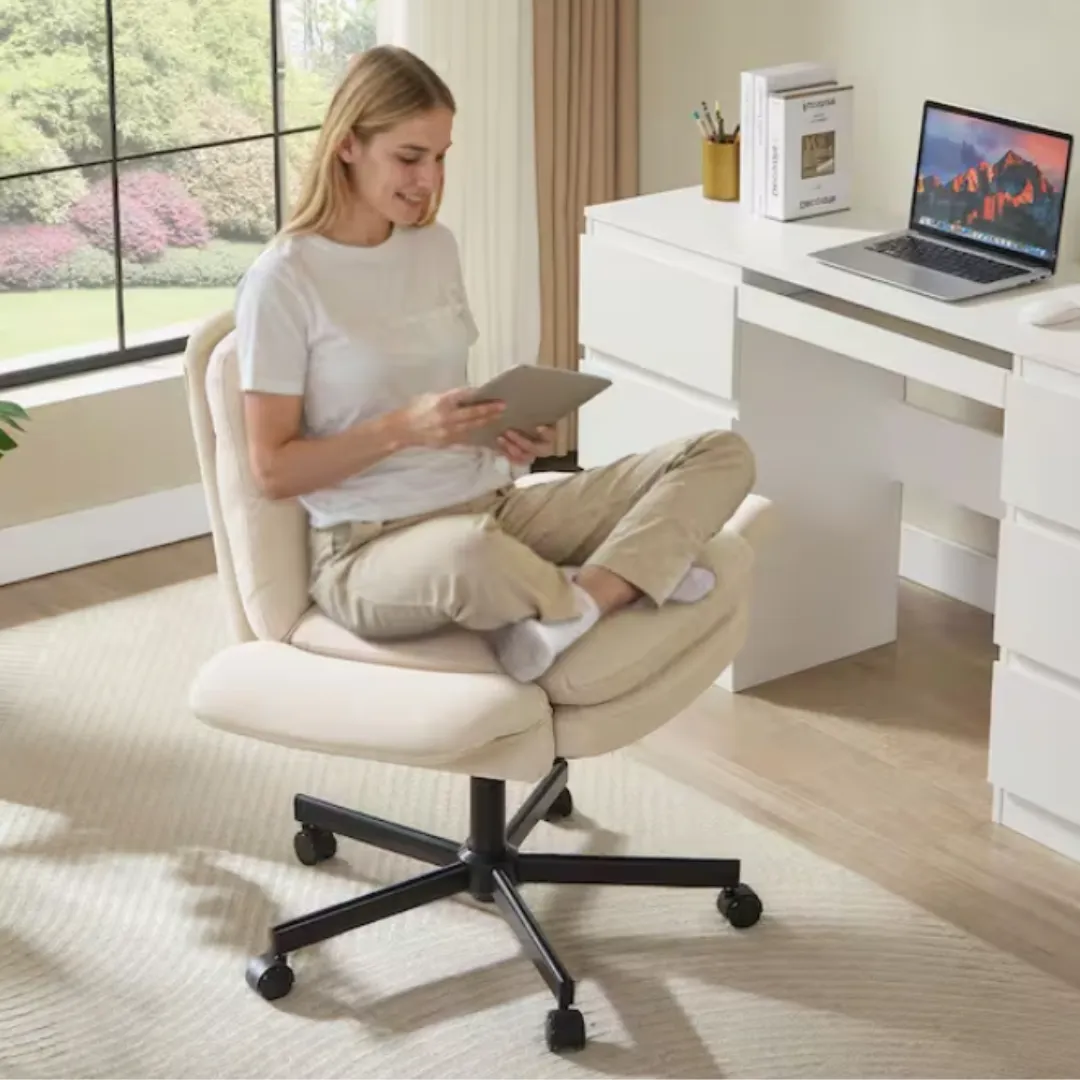 A person sits cross-legged on an adjustable office chair with a cushioned seat and backrest, holding a tablet near a sleek white desk with a laptop, books, and a pencil holder. The room has large windows and light-colored decor.