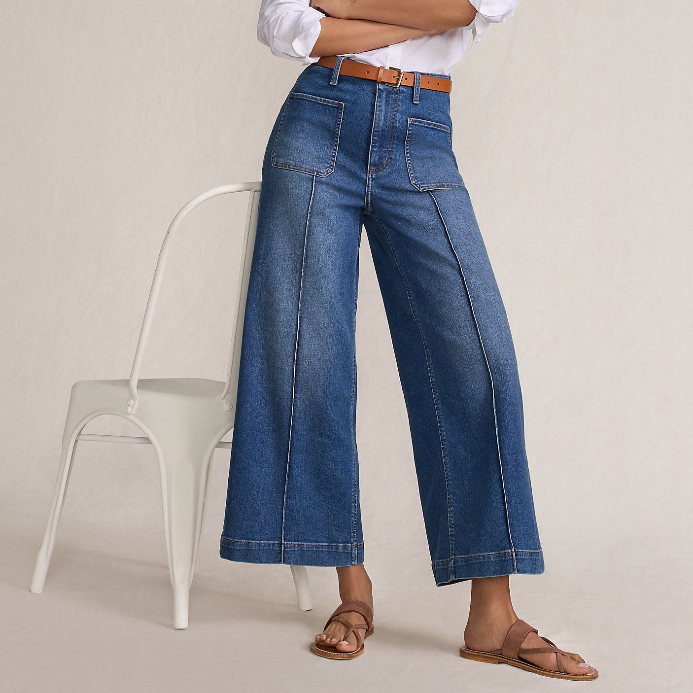 A person is wearing wide-leg denim jeans with patch pockets, a brown belt, a white shirt, and brown sandals, standing near a white chair.