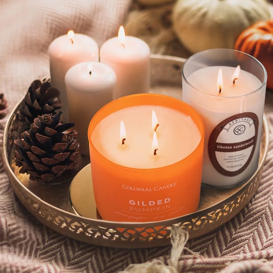 A tray with lit candles including a Colonial Candle \“Gilded Pumpkin\“ and a Heritage \“Tibetan Sandalwood.\“ Pinecones and small pumpkins add a seasonal touch.