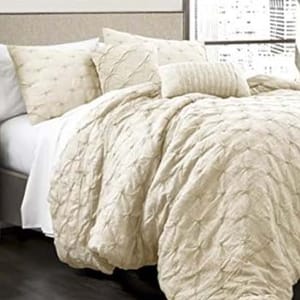 A luxurious bedding set consisting of a textured, cream-colored comforter and several matching pillows, arranged on a bed with a view of a cityscape outside the window.