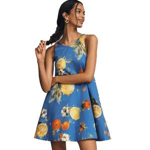 A sleeveless, knee-length dress featuring a blue background with a vibrant fruit and floral print, including lemons and oranges. The model accessorizes with gold hoop earrings.