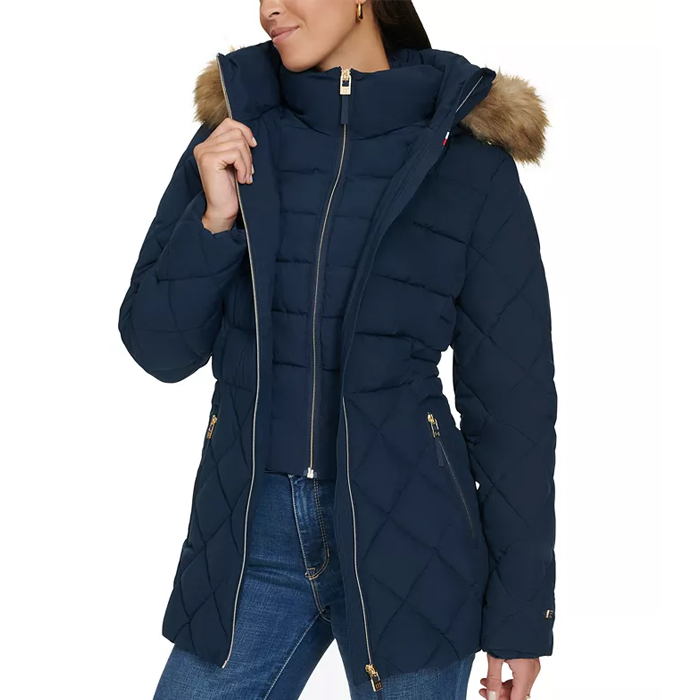 A woman is wearing a navy quilted winter coat with a fur-trimmed hood and a high collar, paired with blue jeans. The coat features a front zipper and zippered pockets.