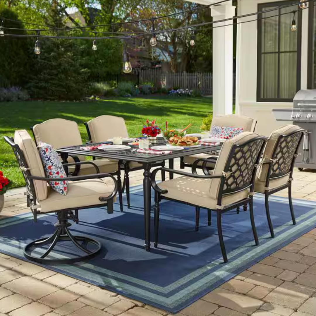 Outdoor patio set featuring a rectangular table with six cushioned chairs, decorative pillows, overhead string lights, and a grill in the background on a blue outdoor rug.