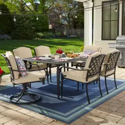 Outdoor patio set featuring a rectangular table with six cushioned chairs, decorative pillows, overhead string lights, and a grill in the background on a blue outdoor rug.
