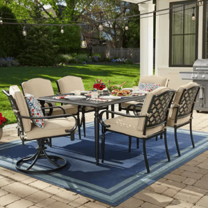 Outdoor patio set featuring a rectangular table with six cushioned chairs, decorative pillows, overhead string lights, and a grill in the background on a blue outdoor rug.