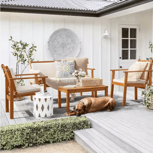 Outdoor patio set featuring two chairs, a loveseat, and a coffee table with beige cushions, set on a deck alongside decorative pillows, a small white side table, plants, and a resting dog.