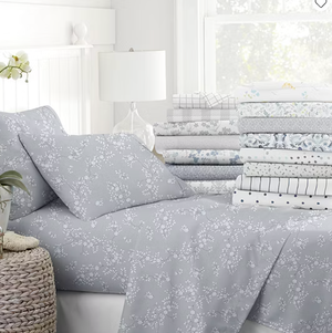 A neatly made bed features a gray floral sheet set alongside a stack of folded sheets with various patterns like plaid, floral, and polka dots, next to a woven basket and glass lamp.