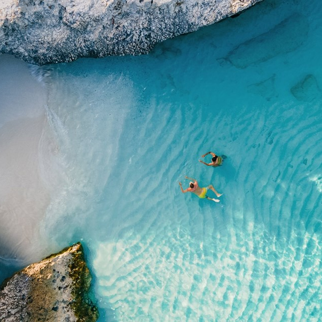 Aerial view of two people swimming in clear turquoise water near a sandy beach and rocky shore.