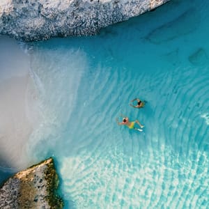 Aerial view of two people swimming in clear turquoise water near a sandy beach and rocky shore.