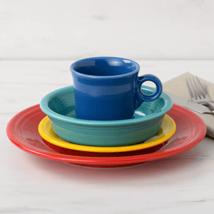A stacked set of colorful dinnerware: a red dinner plate, yellow salad plate, turquoise bowl, and blue mug. A folded napkin with a fork and knife is placed on the side.