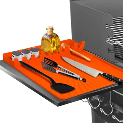 An orange silicone mat on a grill side shelf holds cooking tools, including a spatula, fork, knife, oil bottle, and spice containers.