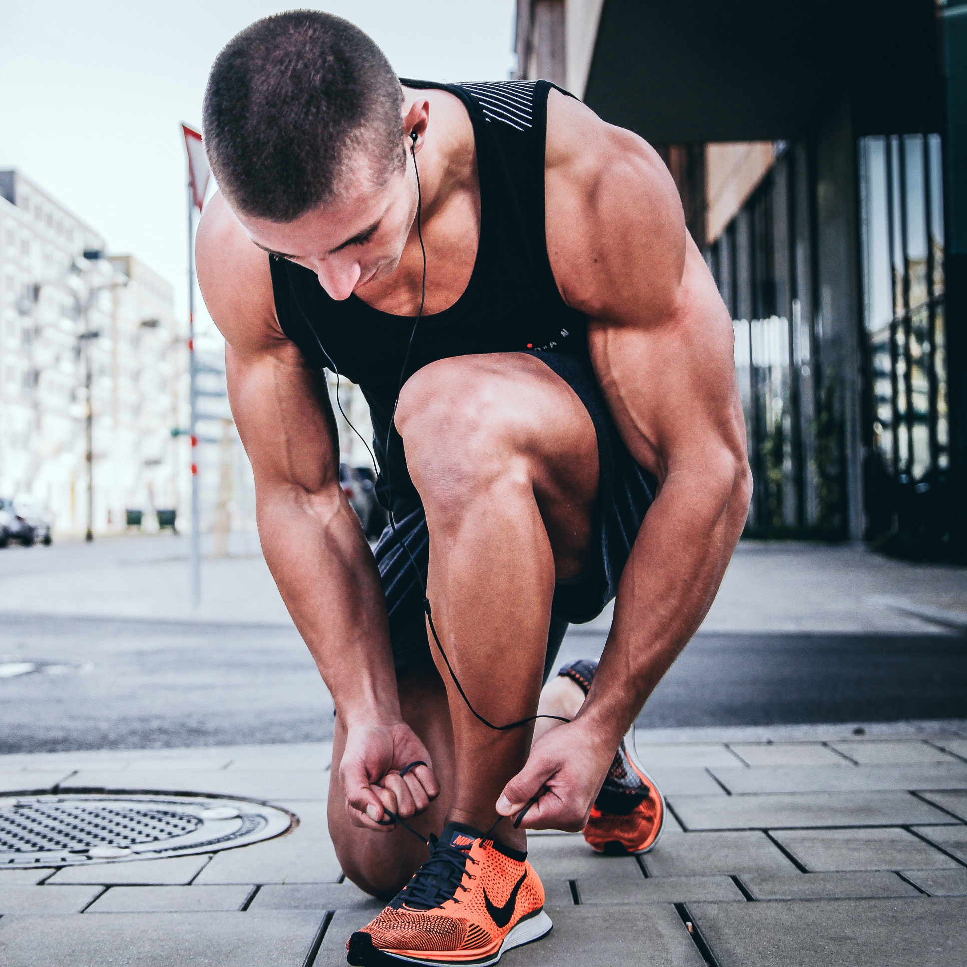 A man tying the laces of bright orange athletic sneakers with a black swoosh logo, wearing a black sleeveless tank top and shorts.