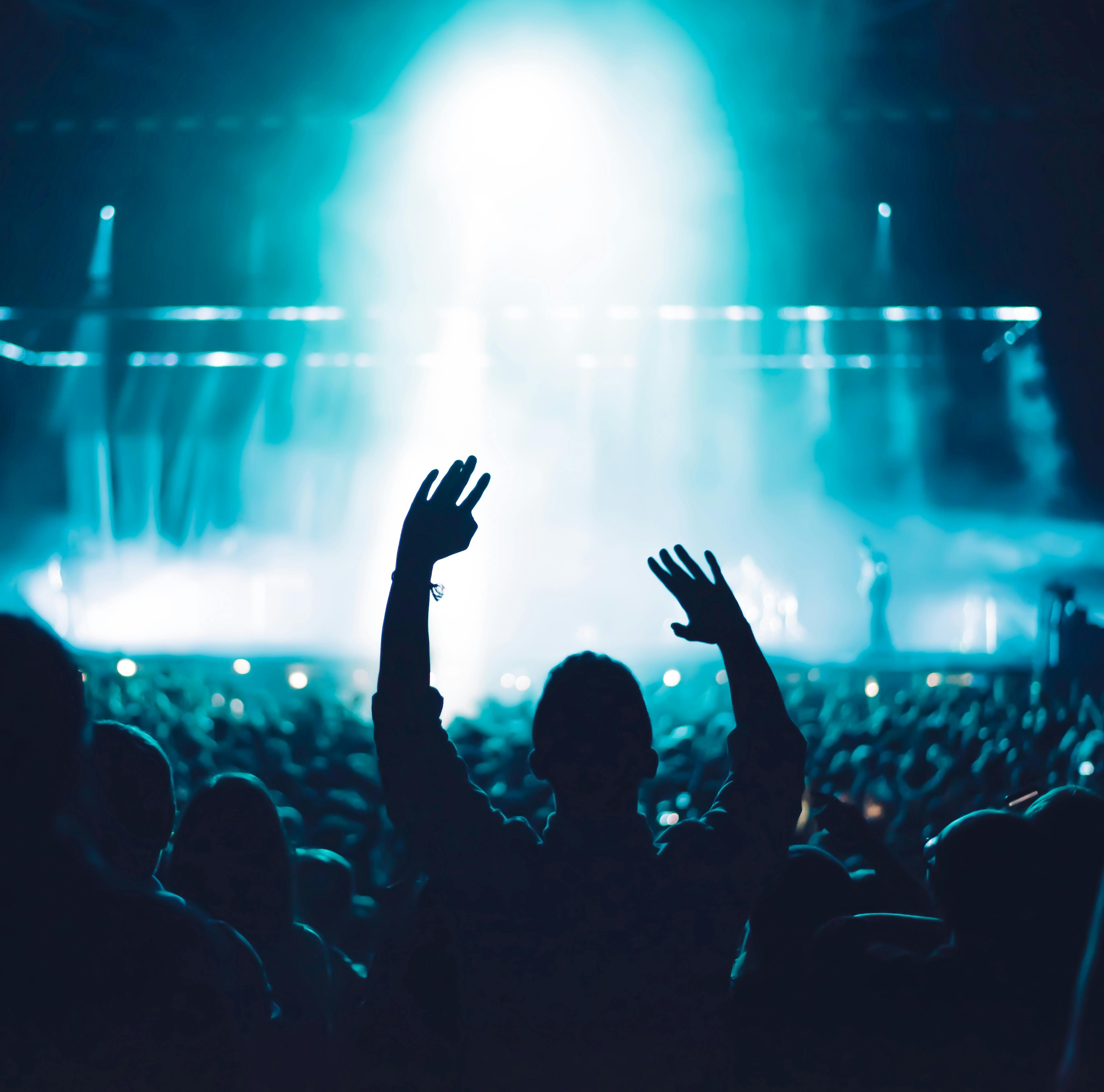 A crowd at a concert with stage lights in the background and a person with raised hands in the foreground.
