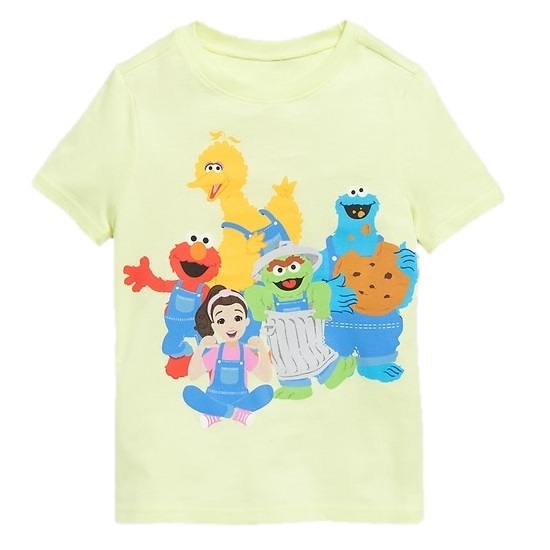 A light yellow t-shirt featuring colorful characters including a yellow bird, red monster, blue monster with cookies, green character in a trash can, and a cartoon girl.