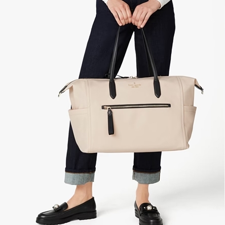 A light-colored handbag held by a person wearing dark loafers, cuffed blue jeans, and holding the bag's black handles.