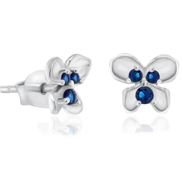 Silver stud earrings shaped like flowers feature three small round blue stones at each center. The petals have a smooth, reflective finish, complementing the vibrant blue gems.