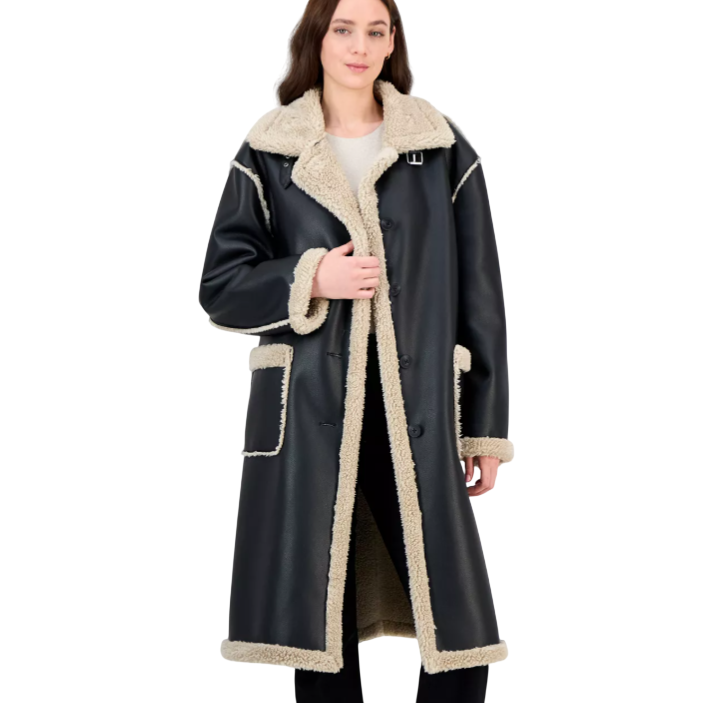 Long black coat with beige shearling trim and lining, featuring a wide collar, large front pockets, and button closure. Styled with black pants and brown shoes.