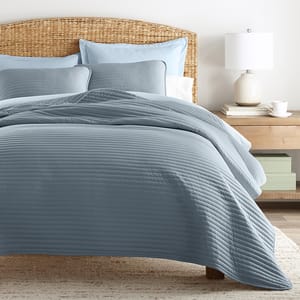 A neatly made bed with blue bedding, including quilted blankets and pillows, sits beside a wooden nightstand with a lamp, a stack of books, and a teacup. The bed has a woven rattan headboard.