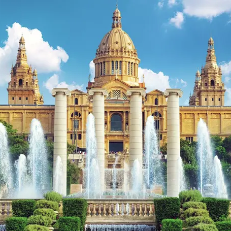 A grand building with twin towers and a central dome is framed by fountains and lush greenery, set against a blue sky with clouds.