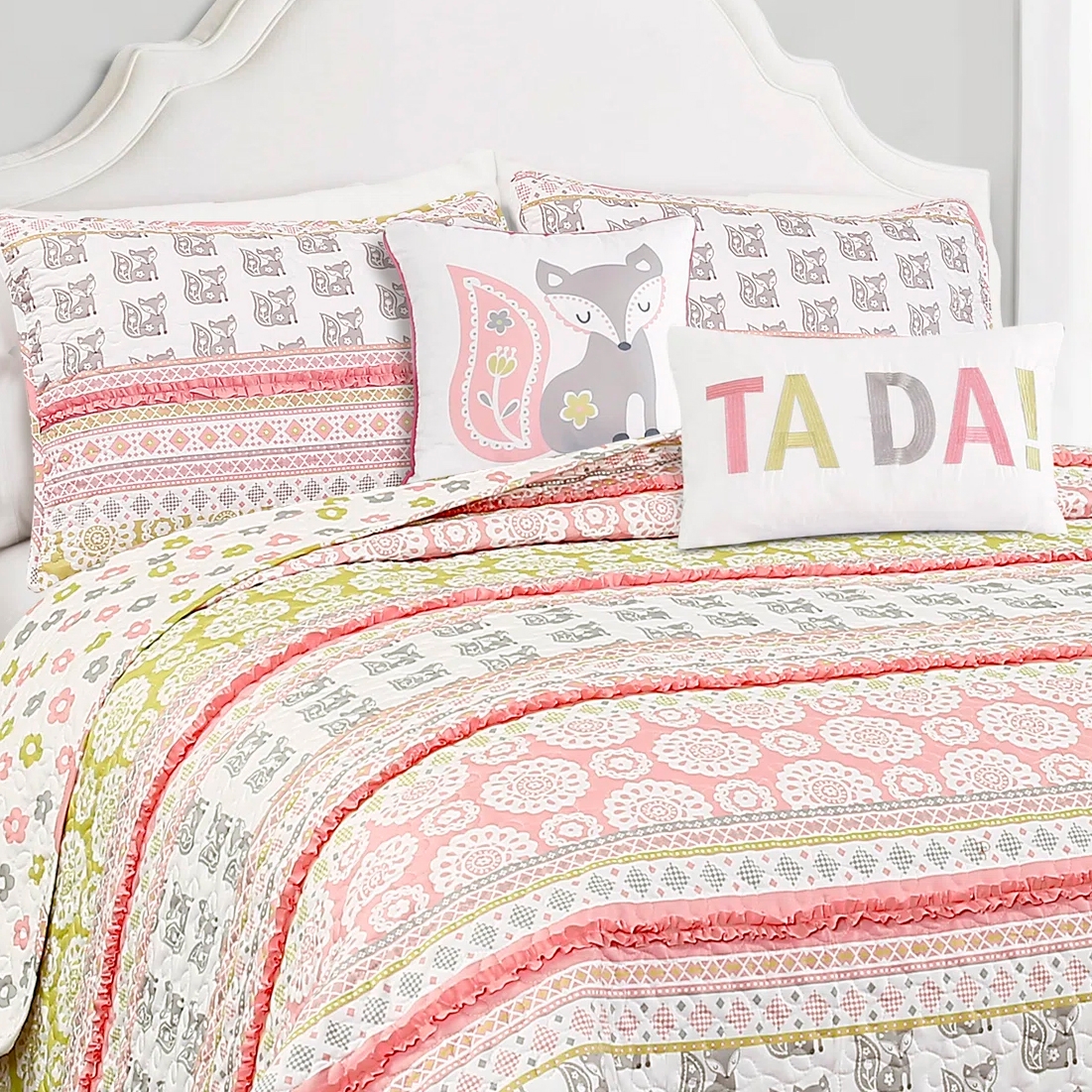 A bedding set featuring a colorful quilt and pillows, including two with a fox design and one that reads \“TA DA!\“ in pastel colors, with patterns in pink, green, and white.
