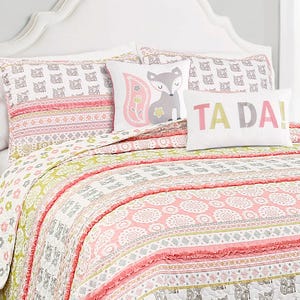 A bedding set featuring a colorful quilt and pillows, including two with a fox design and one that reads \“TA DA!\“ in pastel colors, with patterns in pink, green, and white.