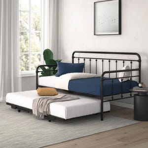 The Wade Logan Beant Daybed with Trundle features a black metal frame with a pull-out trundle for additional sleeping space. It is styled with a navy and white bedding set, complemented by a complementary pillow and throw blanket.
