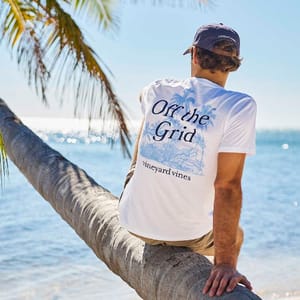 A person sits on a palm tree by the beach wearing a white Vineyard Vines tee with \“Off the Grid\“ on the back, featuring a blue graphic.