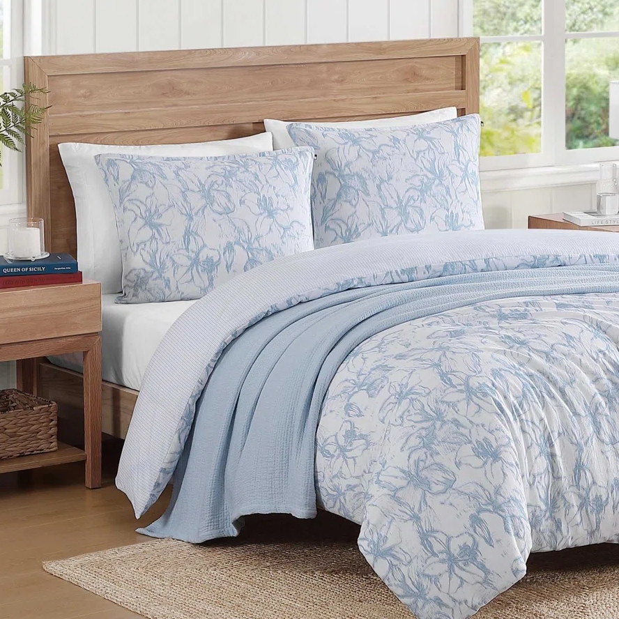 A bedroom setup features a wooden bed with floral-patterned blue and white bedding, including pillows and a comforter. A woven basket and books are displayed on a wooden nightstand, with light streaming in from a nearby window.