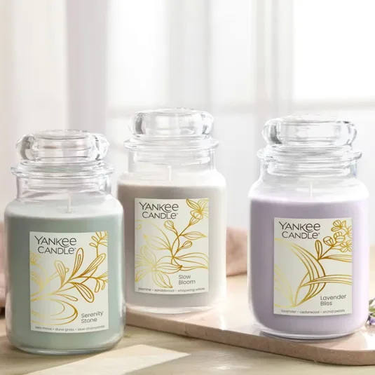 Three Yankee Candle jars are shown: Serenity Stone (sea moss, dune grass, blue chamomile), Slow Bloom (jasmine, sandalwood, whispering willow), and Lavender Bliss (lavender, cedarwood, orchid petals).