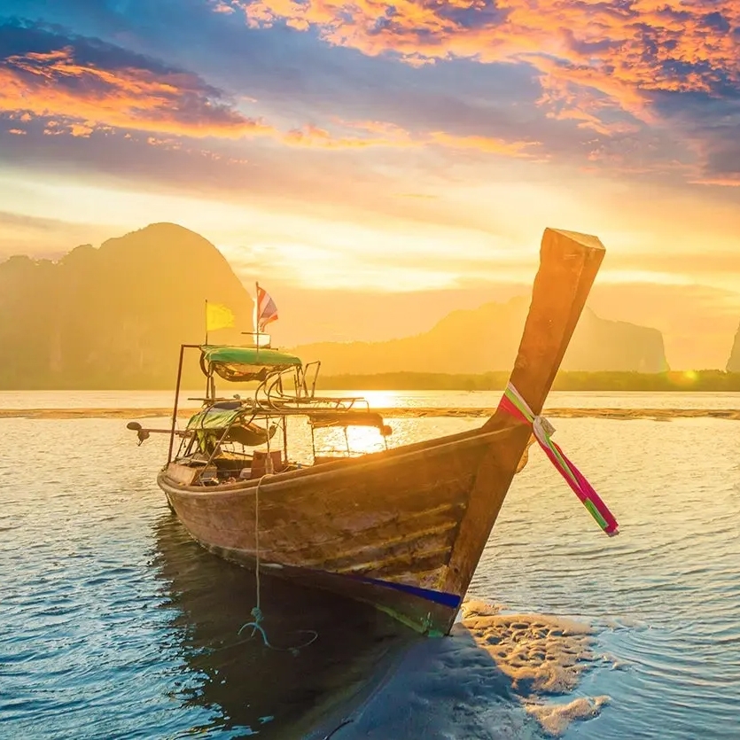 A longtail boat is anchored on a tranquil sea during a vibrant sunset, with mountainous landscapes in the background and a small flag displayed on the boat.