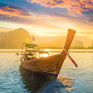 A longtail boat is anchored on a tranquil sea during a vibrant sunset, with mountainous landscapes in the background and a small flag displayed on the boat.