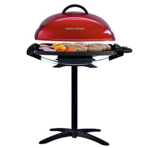Red George Foreman Indoor/Outdoor Grill with a domed lid, cooking vegetables and patties, mounted on a black stand with a wide base for stability.