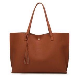 A brown tote bag with a textured surface, featuring two shoulder straps, a tassel detail, and a central button closure.