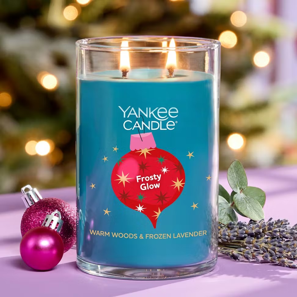 A Yankee Candle labeled \“Frosty Glow\“ with scents of warm woods and frozen lavender is shown lit. It is placed alongside small pink ornaments and sprigs of lavender, with a softly blurred holiday background.