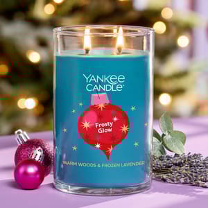 A Yankee Candle labeled \“Frosty Glow\“ with scents of warm woods and frozen lavender is shown lit. It is placed alongside small pink ornaments and sprigs of lavender, with a softly blurred holiday background.