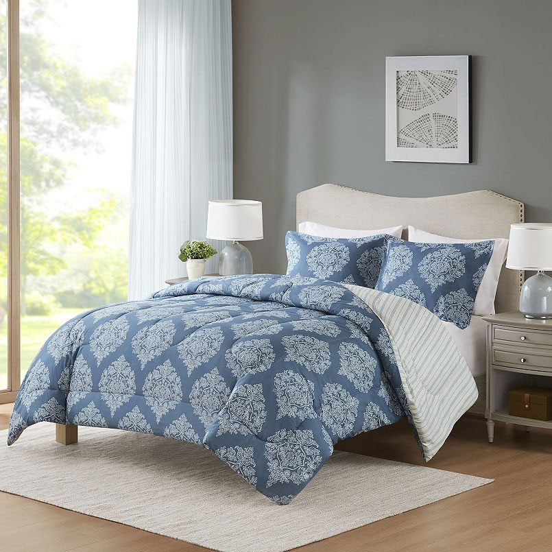 A bedroom setup featuring a blue comforter with white ornate patterns, matching pillowcases, a beige upholstered headboard, and bedside tables with lamps. A large window and framed wall art complete the cozy scene.
