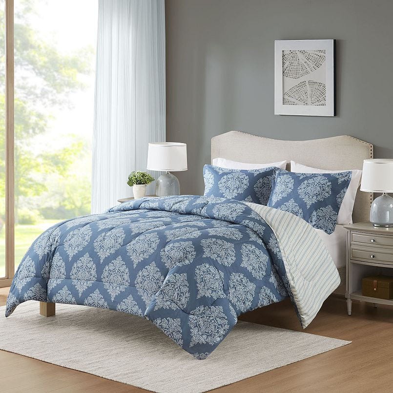 A bedroom setup featuring a blue comforter with white ornate patterns, matching pillowcases, a beige upholstered headboard, and bedside tables with lamps. A large window and framed wall art complete the cozy scene.