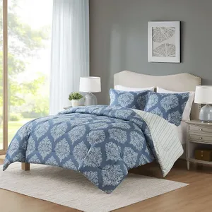 A bedroom setup featuring a blue comforter with white ornate patterns, matching pillowcases, a beige upholstered headboard, and bedside tables with lamps. A large window and framed wall art complete the cozy scene.
