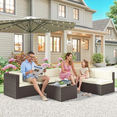 Outdoor furniture set featuring two cream-cushioned brown wicker sofas, a matching square coffee table, and a large umbrella with string lights, placed on a gravel patio in front of a house.