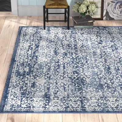 A blue and white patterned area rug is displayed on a light wood floor. A nearby chair, books, and decorative items enhance the setting.