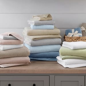A variety of neatly stacked, soft towels in shades of pink, beige, blue, green, and white, displayed on a wooden countertop with some wrapped in a basket.