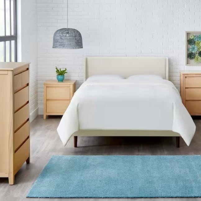 Beige king-sized platform bed with a minimalist design, flanked by two wooden nightstands, set against a white brick wall with a blue rug at the foot.