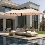 A 13-foot patio umbrella with a beige canopy provides shade over an outdoor sectional sofa next to a pool. The umbrella has a central pole and is set in a modern backyard with large windows, potted plants, and contemporary decor.
