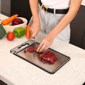 A person cuts raw meat on a metal cutting board, with vegetables like broccoli, carrots, bell peppers, onion, and tomato nearby on a kitchen counter.